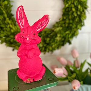 May include: A bright pink ceramic bunny rabbit figurine with white accents on the ears. The bunny is standing on a green metal base.