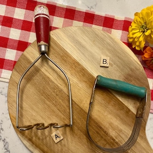 May include: Two vintage kitchen tools on a wooden cutting board. A potato masher with a red and white handle and a pastry blender with a green handle. The cutting board is on a red and white checkered cloth.