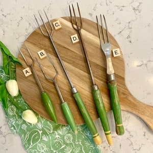 May include: Five vintage green handled metal skewers with wooden handles on a wooden cutting board. The skewers are labeled A, B, C, D, and E.