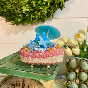 May include: A decorative, pink and white cake-shaped ornament with blue birds, a blue umbrella, and floral accents. The ornament is embellished with sequins and pearls, and sits on a vintage green scale.