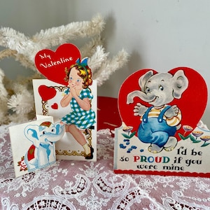 May include: Two vintage Valentine's Day cards featuring cartoon elephants. One card has a red heart with a gray elephant wearing overalls and the text "I'd be so PROUD if you were mine". The other card has a girl in a gingham dress and the text "My Valentine".