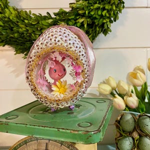 May include: A decorative Easter egg ornament featuring a pink bunny inside, surrounded by pink flowers and gold trim. The egg is adorned with sequins and sits on a vintage green scale, with a green wreath in the background.