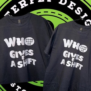 May include: Two black t-shirts with the slogan "WHO GIVES A SHIFT" in white. The design features a gear shift graphic. The shirts are displayed against a green and black background.