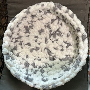 May include: A round, gray and white pet bed with a soft, plush texture. The bed is made of a fuzzy material and has a raised rim.