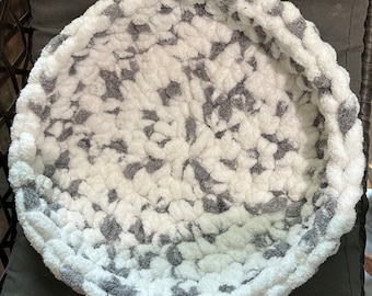 May include: A round, gray and white pet bed with a soft, plush texture. The bed is made of a fuzzy material and has a raised rim.
