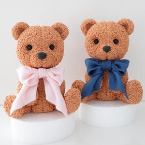 May include: Two brown teddy bear figurines, each with a textured surface, black eyes, and a black nose. One bear has a pink bow, the other a blue bow. They sit on white platforms.