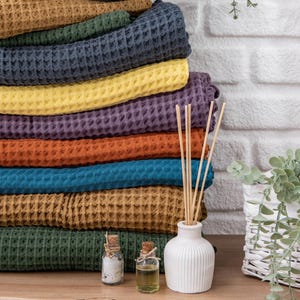 Puede incluir: Una pila de toallas de tejido de gofre en varios colores, incluyendo amarillo mostaza, azul verdoso, naranja óxido y verde oscuro. Las toallas están dobladas y apiladas sobre una superficie de madera. Hay un difusor de cerámica blanca con palitos de madera y dos pequeñas botellas de vidrio con tapones de corcho.