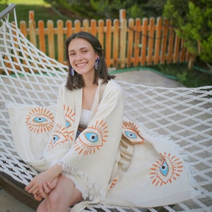 The girl with handmade evil eye kimono poses smiling on the hammock. evil eye kimono is made of 2 layers of muslin and is one hundred percent organic fabric.