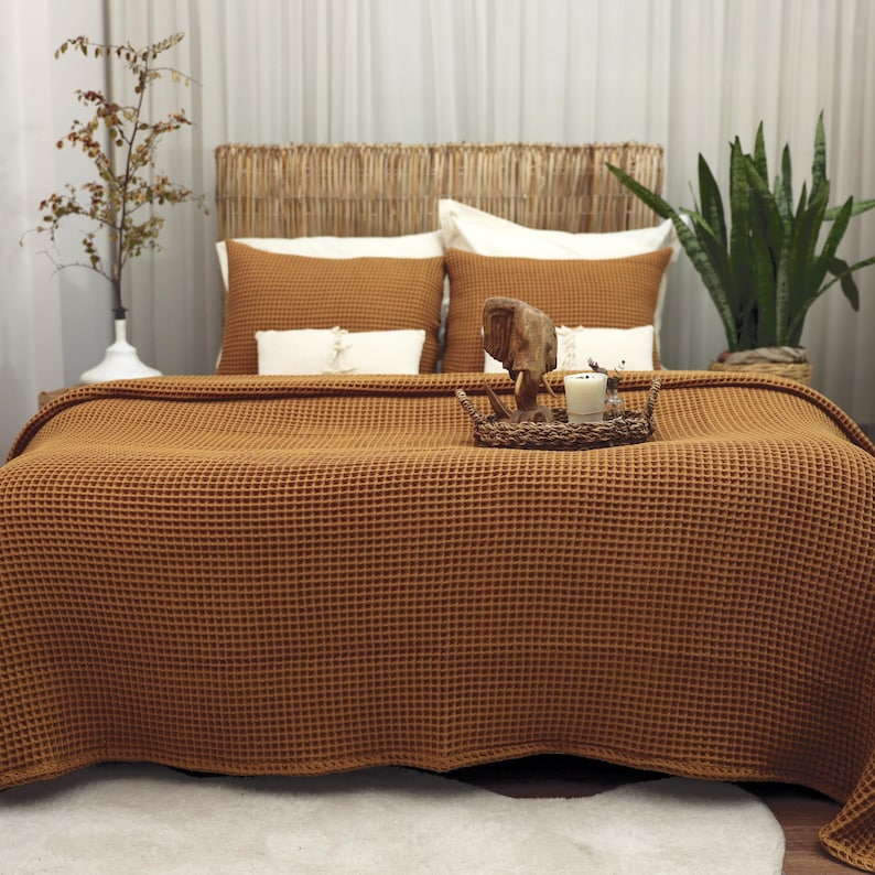 May include: A bed with a woven headboard and a waffle-weave blanket in a warm, earthy tone. The bed is made up with matching pillows and decorative items on a tray, creating a cozy and inviting atmosphere. A potted plant adds a touch of greenery.