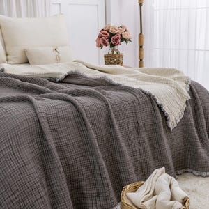 K&ouml;nnte beinhalten: Eine graue Waffel-Decke mit einer wei&szlig;en Waffel-Decke darunter. Die Decken liegen auf einem Bett mit wei&szlig;en Kissen und einem Strau&szlig; rosa Rosen in einem Weidenkorb.