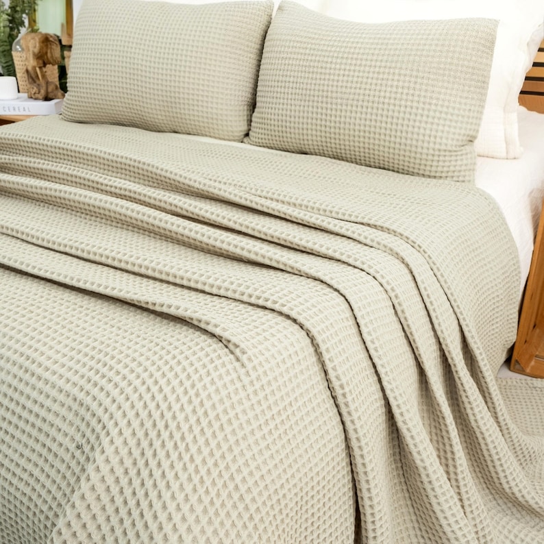 May include: A bed is made with a light beige waffle-weave blanket and two matching pillows. The blanket is draped over the bed, creating soft folds. The pillows are square and have the same textured pattern as the blanket.