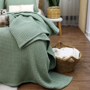 May include: A sage green waffle-weave blanket draped over a bed. The blanket has a textured, grid-like pattern. A woven basket filled with white linens sits nearby. The scene suggests a cozy, comfortable bedroom setting.