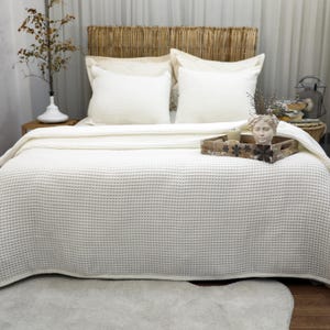 May include: A bed with a woven headboard, two white pillows, and a cream-colored waffle-weave blanket. A wooden tray with a decorative head statue and candles sits on the bed. A vase with branches is on a wooden nightstand.