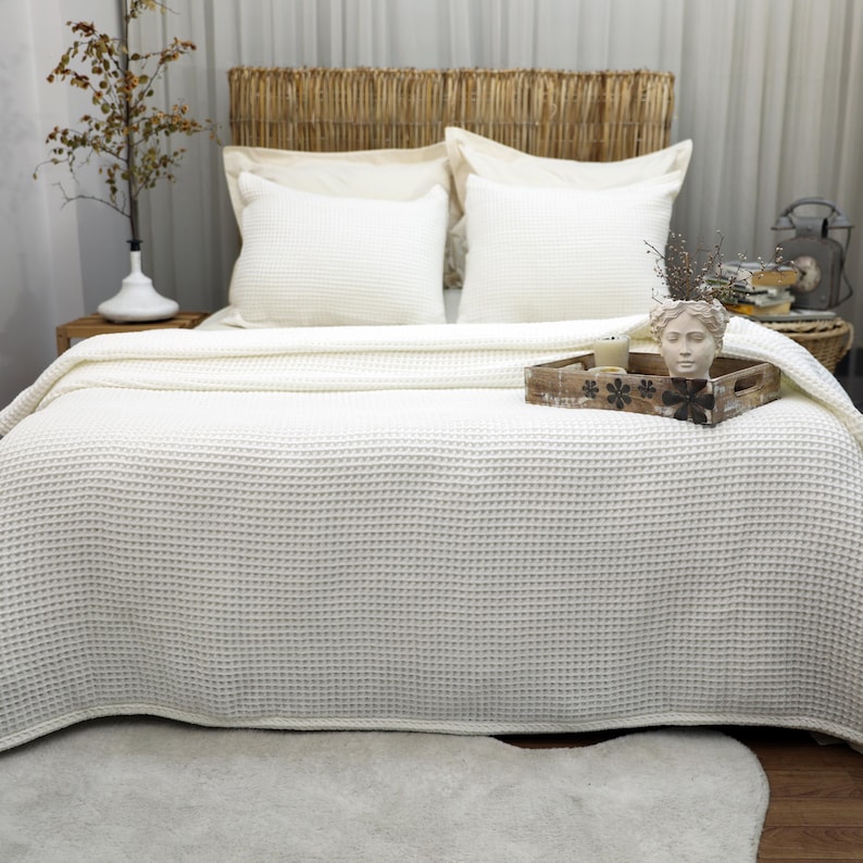 May include: A bed with a woven headboard, two white pillows, and a cream-colored waffle-weave blanket. A wooden tray with a decorative head statue and candles sits on the bed. A vase with branches is on a wooden nightstand.