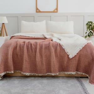 May include: A blush pink and white waffle weave throw blanket with a fringe border. The blanket is folded on a bed with white pillows.