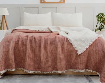 May include: A blush pink and white waffle weave throw blanket with a fringe border. The blanket is folded on a bed with white pillows.