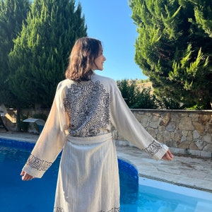 A woman standing in front of a pool and trees with her back to the camera. The model is wearing a beige robe with
black patterns on the back and sleeves
and the bottom hem of the robe is visible above the knees. She is smiling and looking to the side