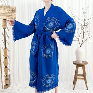 Pode incluir: Robe azul vibrante com desenhos de olhos bordados em fio prateado. O robe apresenta mangas largas, um cinto a condizer e franjas nas bordas. A pe&ccedil;a &eacute; feita de um tecido macio, adequado para relaxar. Mede aproximadamente 140 cm.