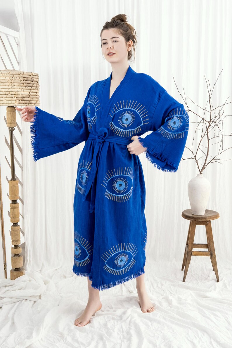 Pode incluir: Robe azul vibrante com desenhos de olhos bordados em fio prateado. O robe apresenta mangas largas, um cinto a condizer e franjas nas bordas. A pe&ccedil;a &eacute; feita de um tecido macio, adequado para relaxar. Mede aproximadamente 140 cm.