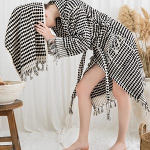 May include: A black and white patterned robe and towel set. The robe has a matching belt and fringe detailing. The towel is draped over the head. The set is made of a soft, textured fabric. The overall theme is spa and relaxation.