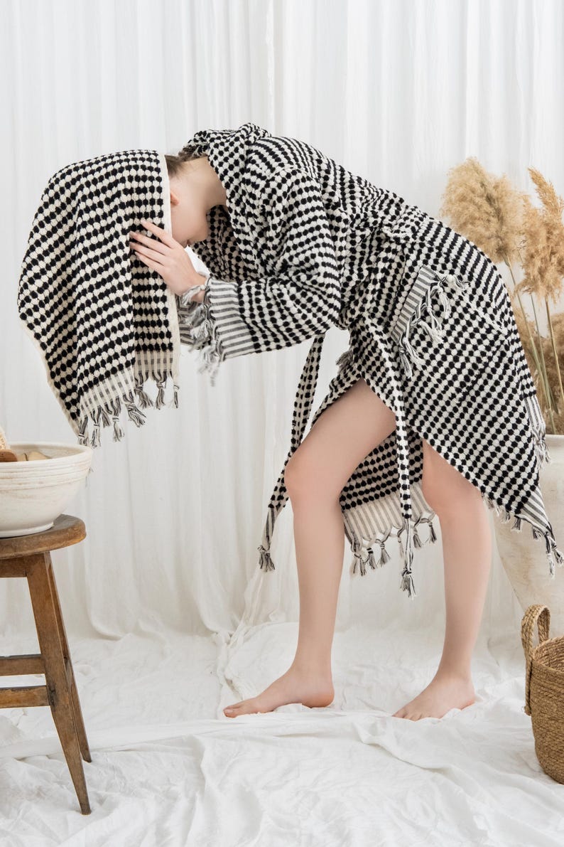 May include: A black and white patterned robe and towel set. The robe has a matching belt and fringe detailing. The towel is draped over the head. The set is made of a soft, textured fabric. The overall theme is spa and relaxation.