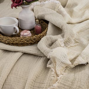 May include: A cream-colored waffle weave throw blanket with a frayed edge. The blanket is folded and draped over a wicker tray with a white vase of pink roses, a small glass bottle, and a pink decorative box.
