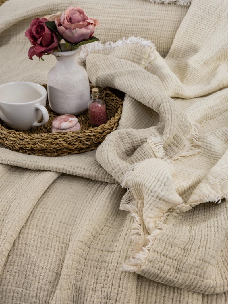 May include: A cream-colored waffle weave throw blanket with a frayed edge. The blanket is folded and draped over a wicker tray with a white vase of pink roses, a small glass bottle, and a pink decorative box.