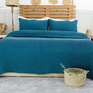 May include: A teal waffle-weave bedspread and two matching pillowcases on a bed with a wooden headboard. The bed is made up with a neutral-colored fitted sheet. A woven basket with greenery sits on the floor.