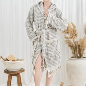 May include: A gray and white patterned hooded bathrobe with a matching belt and fringe trim. The robe is made of a textured fabric and has pockets. A wooden stool holds a bowl with bath accessories.