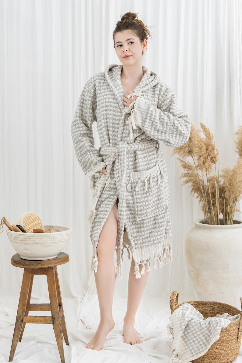 May include: A gray and white patterned hooded bathrobe with a matching belt and fringe trim. The robe is made of a textured fabric and has pockets. A wooden stool holds a bowl with bath accessories.