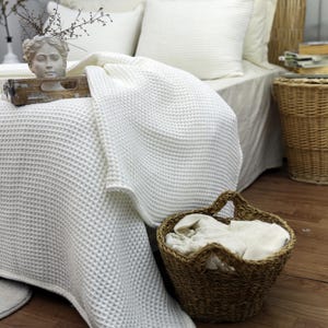 May include: A white waffle-weave blanket draped over a bed, with matching pillows. A decorative statue and a wooden tray are on the bed. A woven basket holds folded linens. The scene evokes a sense of comfort and relaxation.