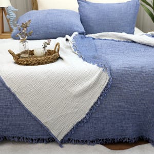 May include: A bed is covered with a blue and white textured blanket, with matching blue pillows. A woven tray holds a candle and small vases with dried flowers. The blanket has a fringed edge.