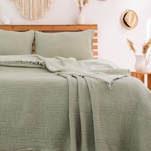 Puede incluir: Una manta de tejido de gofre verde salvia con flecos. La manta est&aacute; doblada en una cama con fundas de almohada a juego.