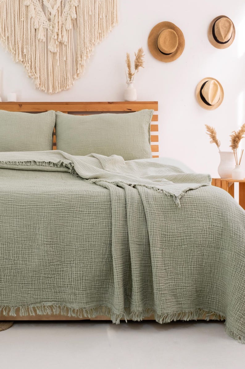 Puede incluir: Una manta de tejido de gofre verde salvia con flecos. La manta est&aacute; doblada en una cama con fundas de almohada a juego.