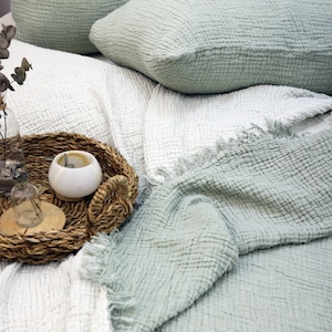 Może przedstawiać: Aranżacja łóżka z białą i jasnozieloną teksturowaną narzutą, z pasującymi poduszkami. Pleciona taca zawiera biały świecznik i małą szklaną butelkę, dodając dekoracyjnego akcentu.
