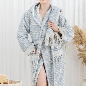 May include: A light blue and white patterned hooded bathrobe with a matching belt and fringed edges. The robe has pockets and is made of a textured fabric. The model is wearing the robe.