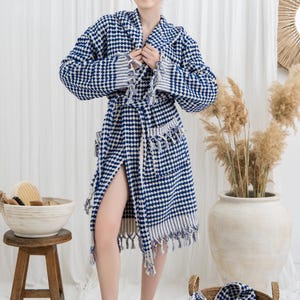 May include: A blue and white checkered robe with a shawl collar and fringe trim. The robe is worn by a person standing in front of a white curtain. A wooden stool and a basket with bath accessories are in the foreground.