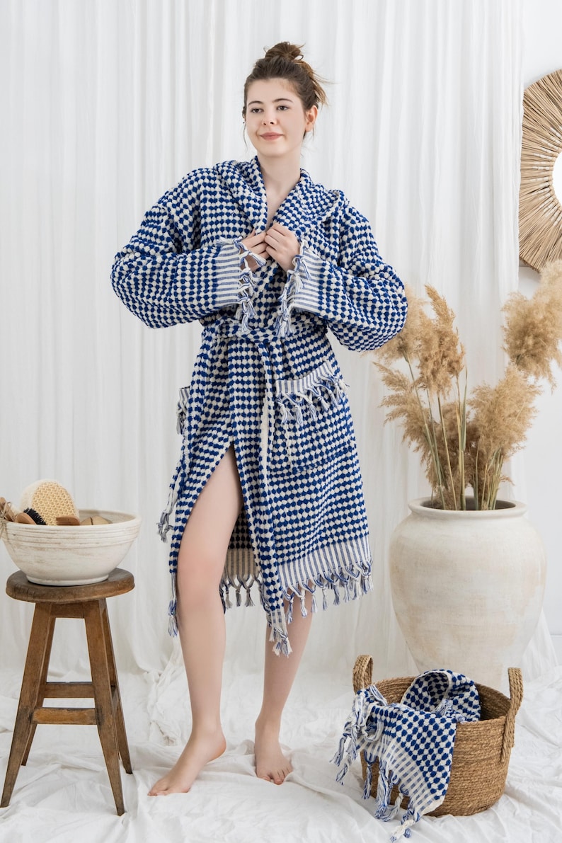 May include: A blue and white checkered robe with a shawl collar and fringe trim. The robe is worn by a person standing in front of a white curtain. A wooden stool and a basket with bath accessories are in the foreground.