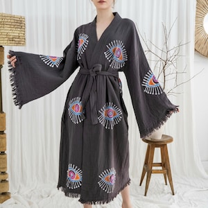 Pode incluir: Robe cinzento escuro com desenhos de olhos bordados em laranja, azul e branco. O robe tem mangas largas com franjas e um cinto a condizer. A modelo est&aacute; a usar o robe em frente a uma cortina branca.