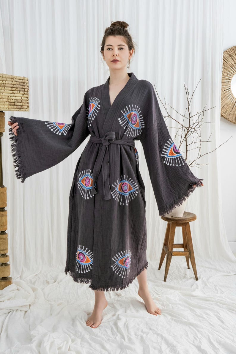 Pode incluir: Robe cinzento escuro com desenhos de olhos bordados em laranja, azul e branco. O robe tem mangas largas com franjas e um cinto a condizer. A modelo est&aacute; a usar o robe em frente a uma cortina branca.