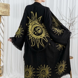 Pode incluir: Robe preta com desenhos dourados de sol e lua. O robe tem mangas compridas com franjas nos punhos e na bainha. Inclui um cinto a condizer. O robe &eacute; feito de um tecido macio e fluido.