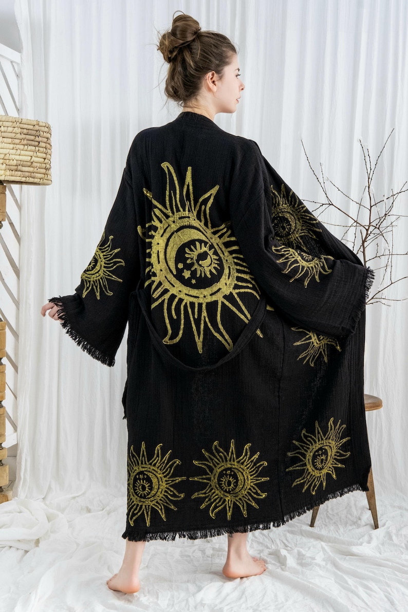 Pode incluir: Robe preta com desenhos dourados de sol e lua. O robe tem mangas compridas com franjas nos punhos e na bainha. Inclui um cinto a condizer. O robe &eacute; feito de um tecido macio e fluido.