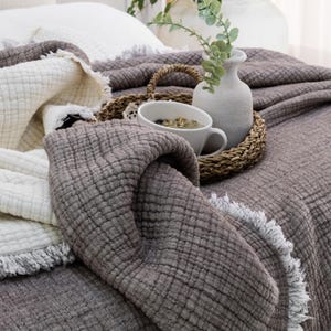 May include: A brown waffle weave throw blanket with a white fringe border. The blanket is folded on a bed with a white pillow and a white throw blanket. A white vase with green leaves and a white mug with tea are on a woven tray.