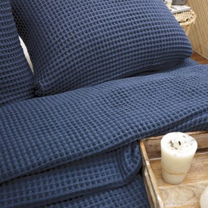 May include: A close-up of a navy blue waffle-weave bedding set, including a pillow and a folded blanket. The textured fabric is visible, and a lit candle sits on a wooden tray in the foreground. The bedding is neatly arranged.