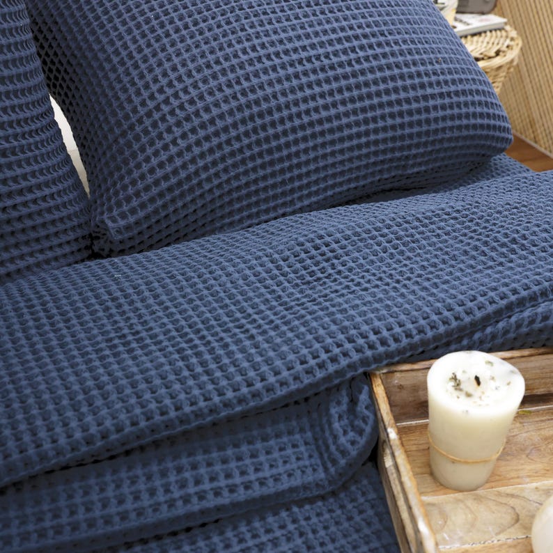 May include: A close-up of a navy blue waffle-weave bedding set, including a pillow and a folded blanket. The textured fabric is visible, and a lit candle sits on a wooden tray in the foreground. The bedding is neatly arranged.