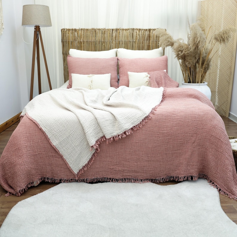 May include: A bed with a woven headboard is covered with a dusty rose comforter and matching pillows. A cream-colored throw blanket with fringe is draped over the comforter. A white rug sits on the floor in front of the bed.