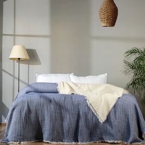 K&ouml;nnte beinhalten: Eine blaue und wei&szlig;e Waffel-Webdecke mit Fransenrand liegt &uuml;ber einem Bett mit wei&szlig;en Kissen. Das Bett steht in einem Raum mit einer Lampe und einer H&auml;ngeleuchte aus Weidengeflecht.