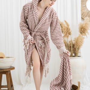 May include: A waffle-weave, mauve and white patterned robe with a matching towel. The robe has a shawl collar, a waist tie, and fringe detailing. The towel is being held over a woven basket.