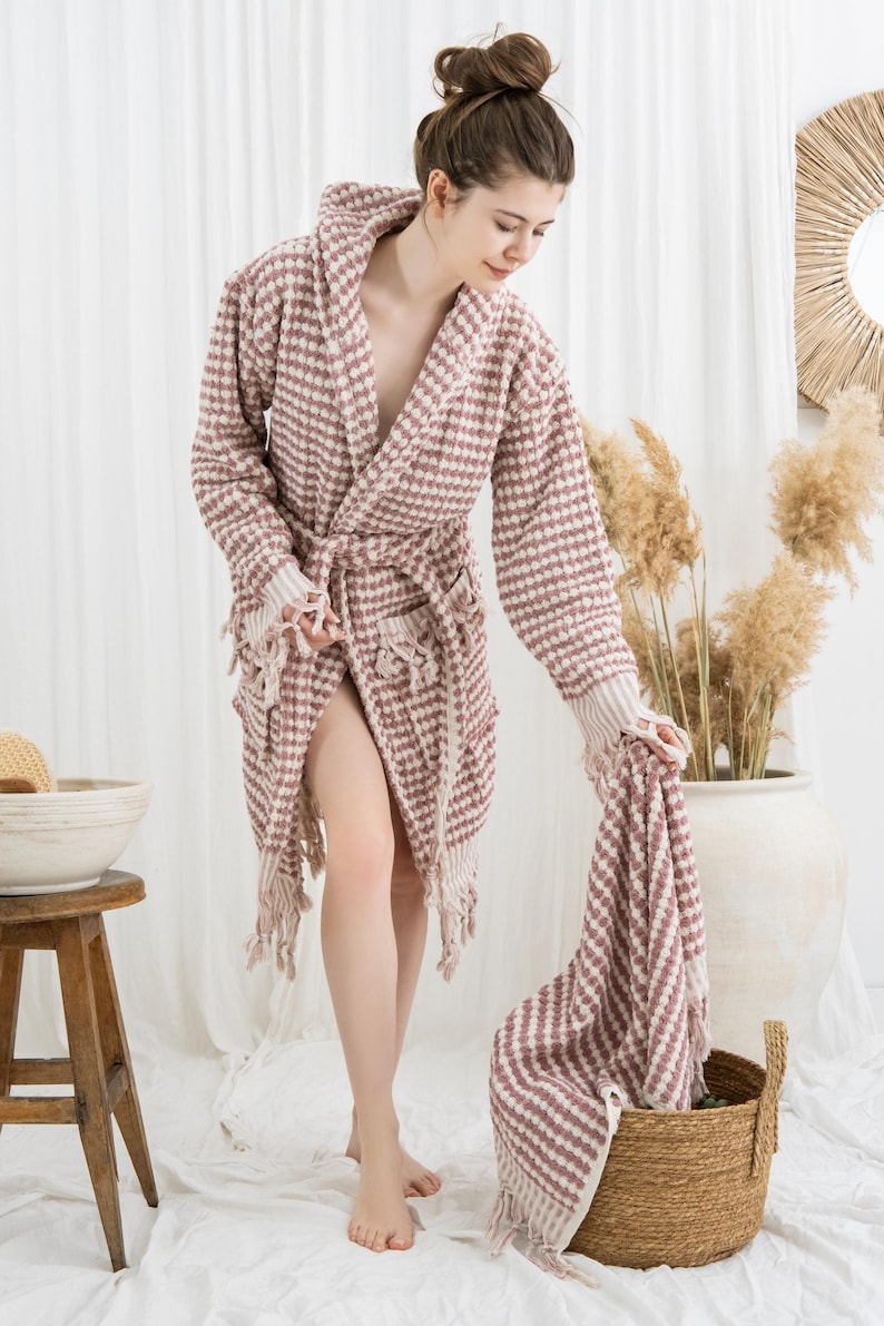 May include: A waffle-weave, mauve and white patterned robe with a matching towel. The robe has a shawl collar, a waist tie, and fringe detailing. The towel is being held over a woven basket.