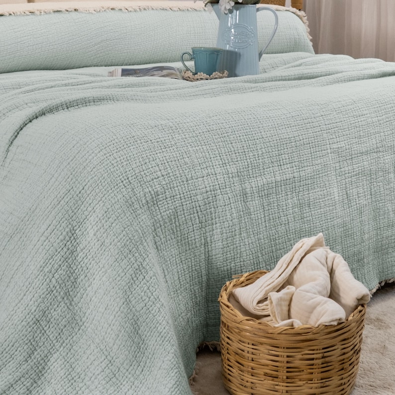 May include: A light green waffle weave throw blanket with a frayed edge. The blanket is folded on a bed with a white pillow and a wicker basket with a white towel.